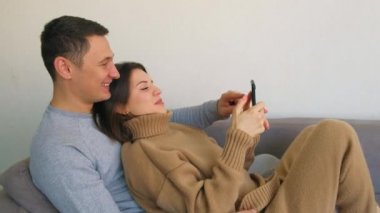 Aşık bir çift, evde kanepede kucaklaşmak, alışveriş için cep telefonu kullanmak, haberleri okumak ve aile veya akrabalarla mesajlaşmak. Akıllı bir telefona bakarak neşeli bir iletişim. Birbirine aşık çift.