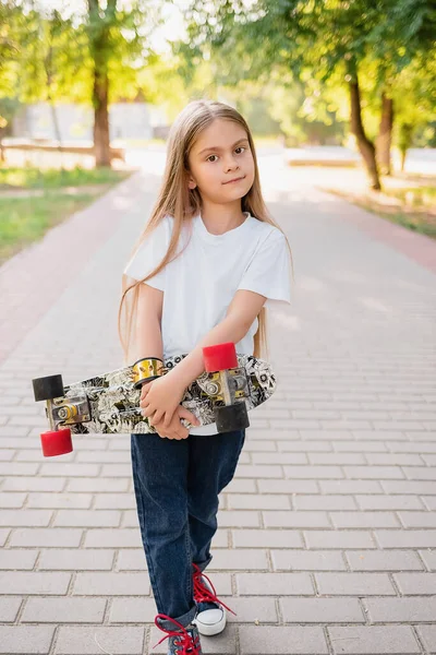 Parkta günlük giysiler içinde güzel bir beyaz kız öğrencinin portresi. Elinde bir kaykay tutuyordu. Beyaz tişört ve kot pantolon giymişti. Çocuk giyimi reklamcılığı konsepti.