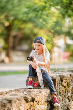 Beyzbol şapkalı küçük bir kız bir taşın üzerinde ters oturuyor, elleri dizinde kaykayı tutuyor, bir bacağı sarkıyor..
