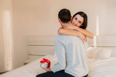 Mutlu genç kadın elinde hediyeyle kocasına sarılıyor. Olumlu duygular. Yatak odasında sevgi dolu bir çift. Gençlerin günlük yaşamları.