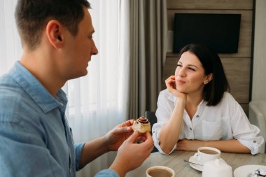 Young beautiful guy and girl at home in living room have breakfast in front of window. Girl admiring a guy listens to him attentively and smiles. Home atmosphere. Camera focus on the girl