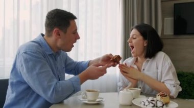 Genç çift mutfakta kahvaltı yaparken eğleniyor. Adam kadını pastayla besliyor. Gençler sabahları eğleniyor. Mutlu aile.