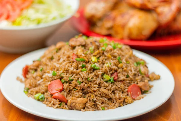 Chaufa rice in a white plate, on a wooden table, with an out of focus background grilled chicken and salad.