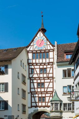 Eski koyu kahverengi ortaçağ saat kulesi. Güneşli bir günde pastel renkli ve altın yuvarlak saati olan beyaz, yarı ahşap bir apartmanda tatlı rüzgar gülü.