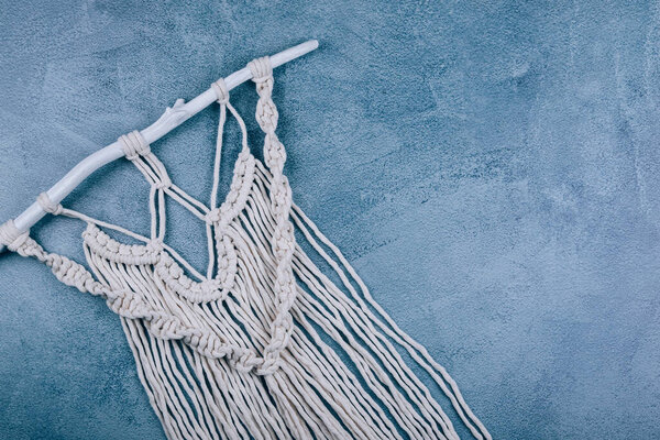 Handmade wall decoration cotton macrame panel on wooden stick over textured blue gray background.