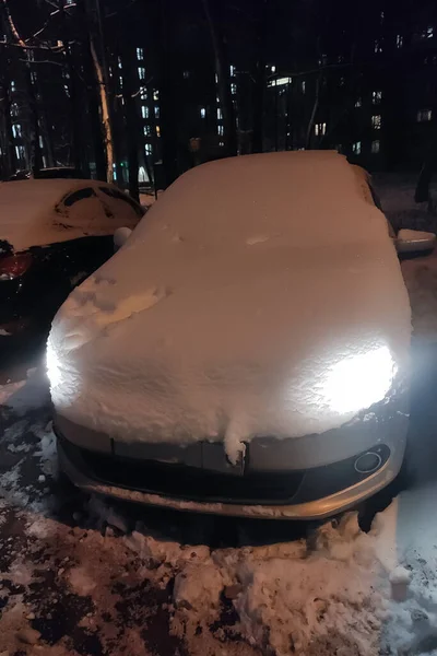 車は夕方に雪の厚い層で覆われています この車のヘッドライトがオンになって雪の層を通して輝きます — ストック写真