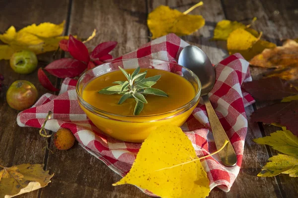 秋のカラフルな葉と小さな野生のリンゴで飾られた木製のテーブルの上にガラスボウルにカボチャのスープ — ストック写真