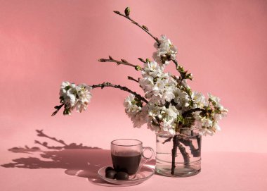 Cam bir vazonun yanında şeffaf bir fincan kahve ve pembe arka planda çiçek açan bahar dalları.