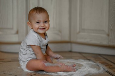Küçük bir çocuk un kaplı mutfakta oturuyor ve un ile oynuyor.