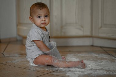 Küçük bir çocuk un kaplı mutfakta oturuyor ve un ile oynuyor.