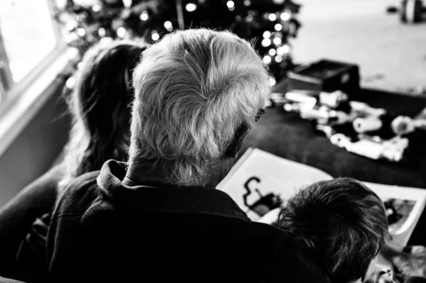 Дедушка читает сказку двум внукам, сидящим у него на коленях — стоковое фото