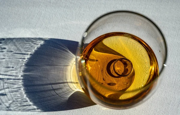 Light refracting through a glass of amber colored liquid with a wedding ring at the bottom — Stock Photo, Image