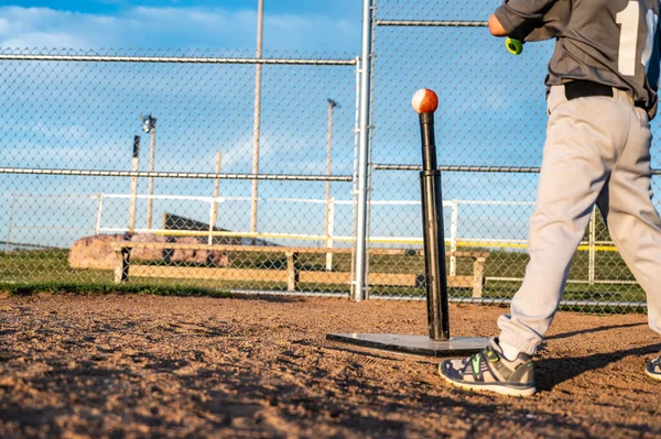 Jovem jogador de beisebol em pé no home plate para bater uma bola fora de um tee com um taco. — Fotografia de Stock