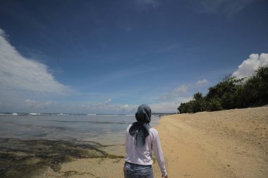 Tesettürlü ve beyaz tişörtlü Asyalı bir kızın arkası sabah açık mavi gökyüzü ve kabarık bulutlarla kumsalda yürüyor. Seyahat, tatil ve yaz konsepti.