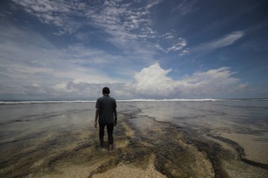 Çıplak ayaklı Asyalı bir adamın arka görüntüsü sabah plajda berrak mavi bir gökyüzü ve tüylü bulutlarla yürüyor. Seyahat, tatil ve yaz konsepti.