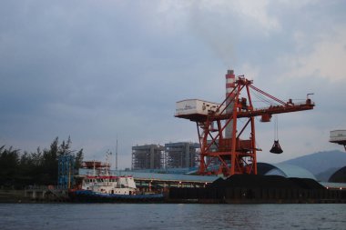 Sukabumi, Batı Java, Endonezya - 23 Ağustos 2021: Kömür ateşiyle çalışan buhar santralinin manzarası kanat vinci ile Pelabuhan Ratu kıyısı yakınlarındaki gemi kargosundan kömür stoklarını kaldırıyor..