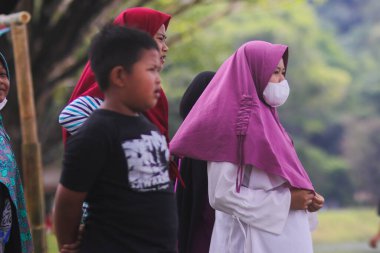 Sukabumi, Batı Java, Endonezya - 19 Ağustos 2021: Mor tesettürlü Endonezyalı bir çocuk salgının yayılmasını önlemek için beyaz bir maske takıyor.