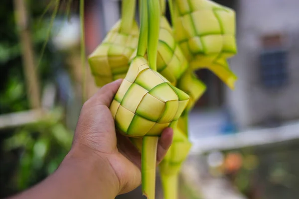 Hand holding a Ketupat or Kupat with blurred background. Ketupat is an iconic rice cake that usually served during Hari Raya or eid mubarak. Ketupat pouch is made of woven young coconut leaves