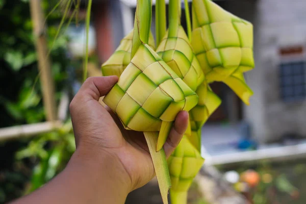 Hand holding a Ketupat or Kupat with blurred background. Ketupat is an iconic rice cake that usually served during Hari Raya or eid mubarak. Ketupat pouch is made of woven young coconut leaves