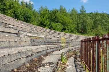 Stadyumun terk edilmiş beton tribünleri, paslı çitlerle çevrili. Zamanın tahribatıyla ısırılmış bir sahne.