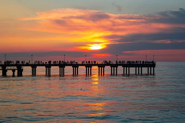 Palanga rıhtımında deniz batımının arka planında dinlenen insanlar var.