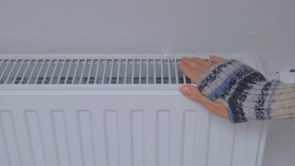 Жінка нагріває руки на опалювальному радіаторі біля стіни. Холод у квартирі — стокове відео