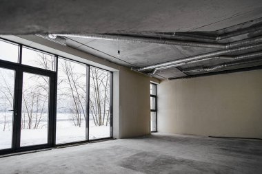 Odanın inşaatı devam ediyor. Duvarları alçılı, zemini beton. Beton bir tavanda havalandırma boruları ve inşaat halindeki bir binada panoramik pencereler..