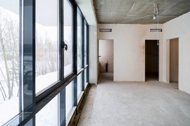 Odanın inşaatı devam ediyor. Duvarları alçılı, zemini beton. Beton bir tavanda havalandırma boruları ve inşaat halindeki bir binada panoramik pencereler..