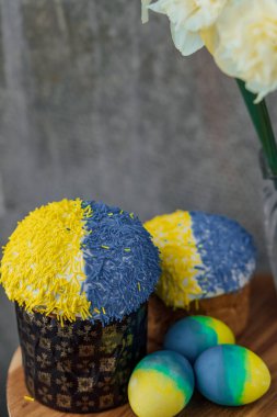 Ukrayna bayrağının renginde lezzetli Paskalya pastaları, arka planda çiçekler olan ahşap bir masada sarı-mavi Paskalya yumurtaları. mesaj için yer. seçici odak