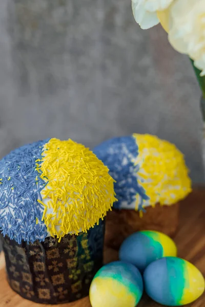 Ukrayna bayrağının renginde lezzetli Paskalya pastaları, arka planda çiçekler olan ahşap bir masada sarı-mavi Paskalya yumurtaları. mesaj için yer. seçici odak