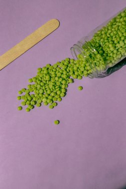 Tüy dökümü için güzel yeşil balmumu tanecikleri açık mor arka planda şeffaf bir kavanozdan, ahşap bir spatuladan dökülür. Epilasyon, tüy dökülmesi, istenmeyen kılların alınması. Yukarıdan görüntüle
