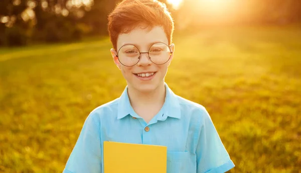 Vidám, stílusos fiú áll a parkban másoló könyvvel a kezében és mosolyog — Stock Fotó
