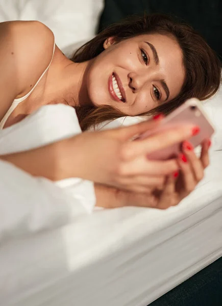 Örömteli fiatal nő használ okostelefon, miközben feküdt az ágyban reggel — Stock Fotó