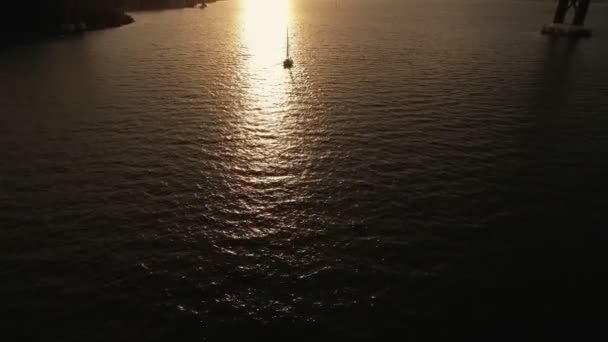 Silhouet van eenzame zeiljacht gaan in de zonsondergang op de rivier onder een brug — Stockvideo