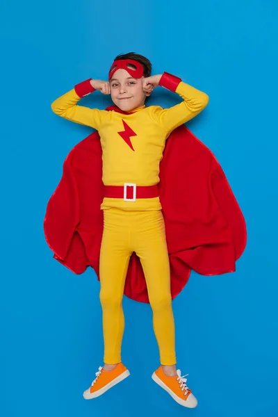 Smiling boy in hero costume lying on blue background — Stock Photo, Image