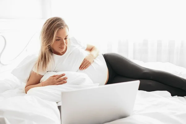 Νεαρή έγκυος γυναίκα που βλέπει φιλμ στο λάπτοπ ξαπλωμένη στο κρεβάτι — Φωτογραφία Αρχείου