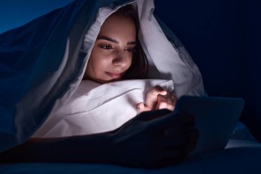 Geceleri kadın battaniyenin altında tablet kullanıyor.