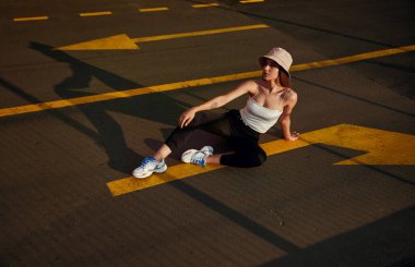 Günbatımında oklarla asfalt yolda otururken şapkalı, modaya uygun giyinmiş yüksek açılı genç bir kadın.