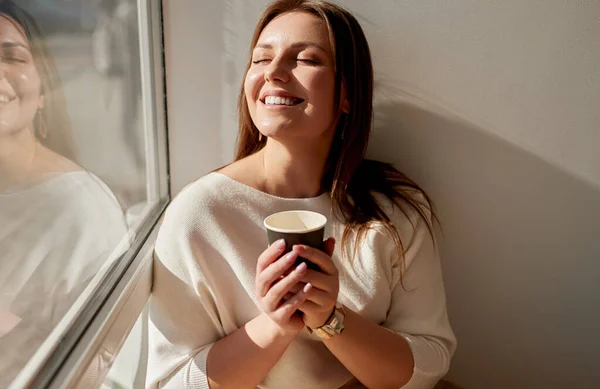 커피를 마시면서 눈을 감고 창문 근처에서 웃고 있는 행복 한 젊은 여성 — 스톡 사진