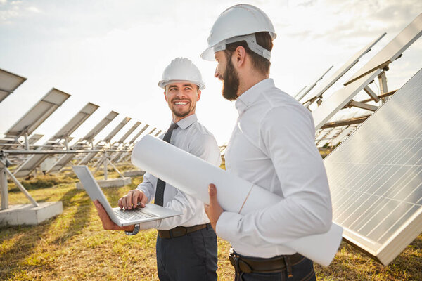 Улыбающиеся предприниматели с ноутбуком и чертежами на солнечной электростанции