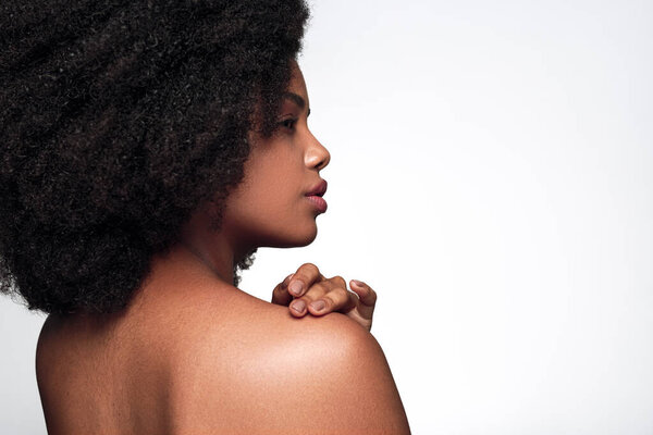 Side view of confident young African American female model with curly dark hair touching bare shoulder and looking away against white background