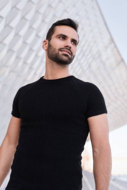 Gündüz vakti modern geometrik binanın yanında dikilirken siyah tişörtlü, gülümseyen ve başka yöne bakan kendine güvenen genç, sakallı bir Arap.