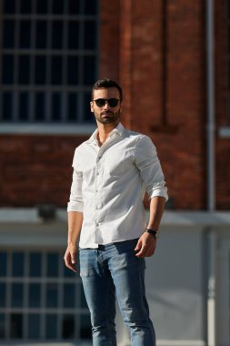 Kendinden emin, siyah saçlı, beyaz tişörtlü ve güneş gözlüklü genç bir adam güneşli bir günde tuğla binanın yanındaki sokakta dikiliyor.