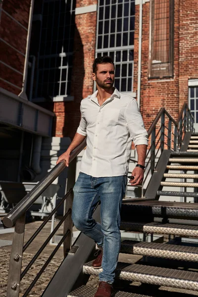 Ethnic bearded male in jeans and white shirt walking down metal stairs and looking away on sunny day outside brick building