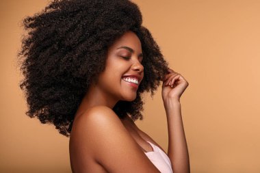 Pozitif özgüvenli genç Afro-Amerikan kadın modelinin kıvırcık siyah saçlara dokunması ve bej arka planda kapalı gözlerle gülümsemesi.