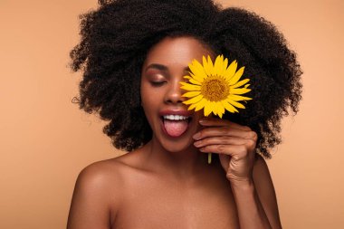 Çıplak omuzları ve kıvırcık saçları olan genç siyah kadın kahverengi arka planda cilt bakımı sırasında gözlerini doğal ayçiçeğiyle kapatıyor.