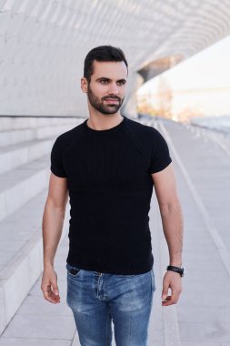 Çatı altında merdivenlerde dikilip gündüz vakti şehir sokağına bakan sıradan giysili yakışıklı bir etnik adam.