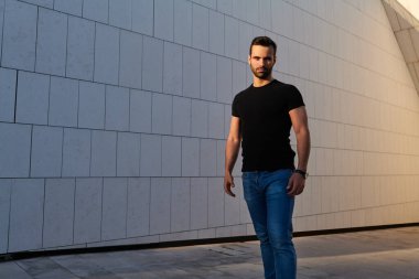 Siyah tişörtlü, esmer ve sakallı, modern binanın yanında asfalt caddede duran ve kameraya özgüvenle bakan ciddi bir Arap erkek.