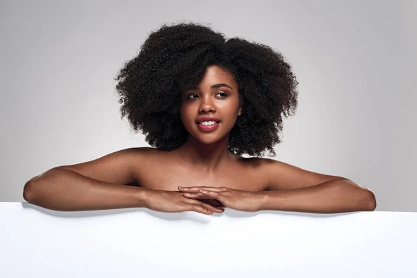 Positive African American female model with bare shoulders and curly hair folding arms on empty white poster during advertisement campaign against gray background