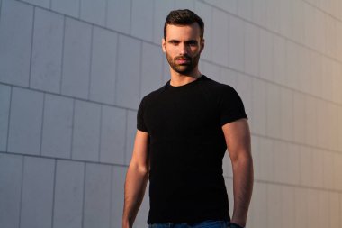 Siyah tişörtlü, koyu renk saçlı ve sakallı, modern binanın yanındaki caddede duran ve kameraya bakan kendinden emin genç, etnik atletik bir erkek.